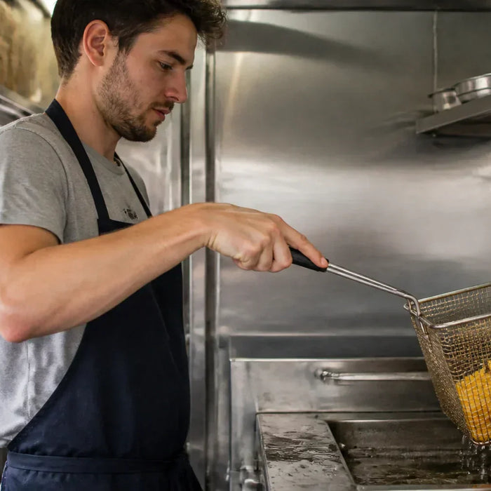 A person in a casual t-shirt and apron working a commercial deep fryer inside a compact food truck kitchen.