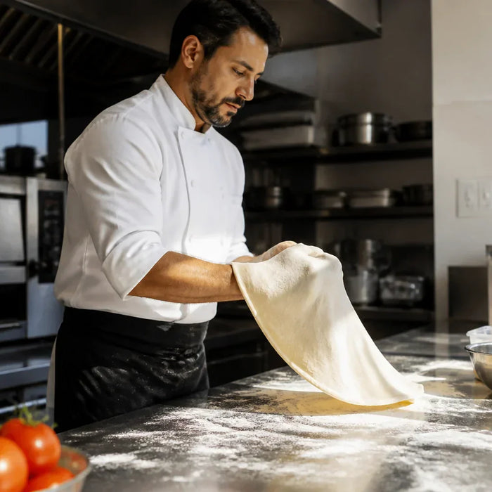 A professional chef in a clean, modern commercial kitchen environment, expertly stretching pizza dough on a large stainless steel prep station.