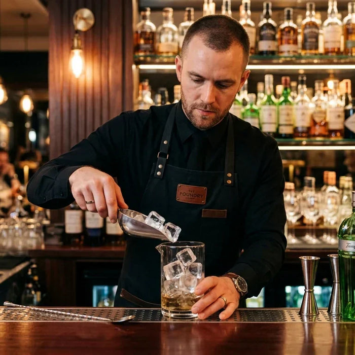 A professional bartender in a clean uniform, focused on crafting a cocktail in a high-end bar setting. They are using a stainless steel scoop to add fresh, crystal-clear ice cubes to a cocktail shaker or a mixing glass