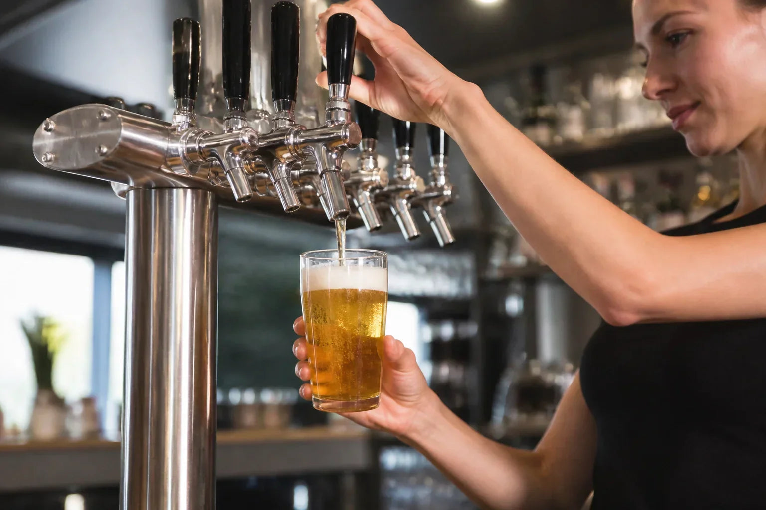 Bartender pouring a draft beer from a stainless steel multi-tap kegerator tower in a modern commercial bar setting.