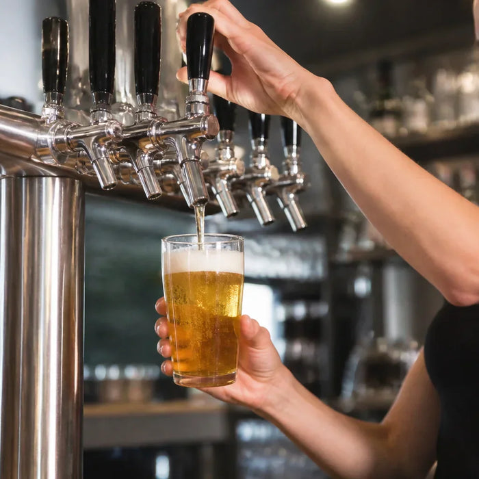 Bartender pouring a draft beer from a stainless steel multi-tap kegerator tower in a modern commercial bar setting.