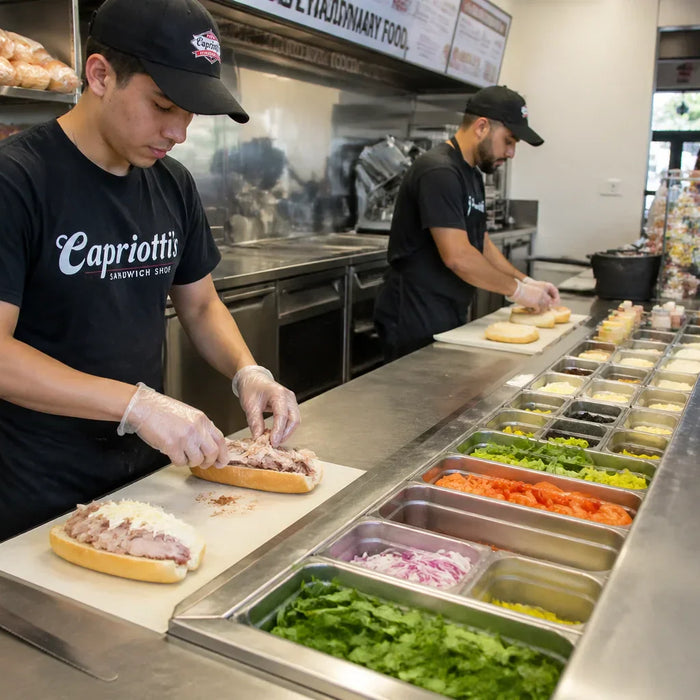 A high-angle shot of a busy sandwich shop during a lunch rush, focusing on a mega top sandwich prep table. 