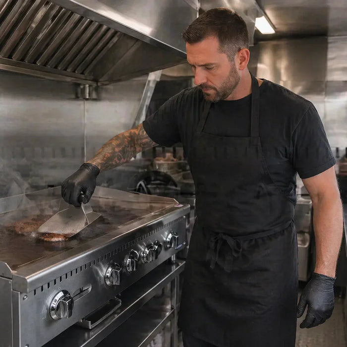 A professional chef in a food truck kitchen smashing a burger on a heavy-duty stainless steel propane griddle
