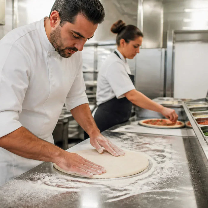 Professional pizza maker stretching dough on a commercial Atosa pizza prep table with chilled topping pans of mozzarella, pepperoni, and fresh vegetables