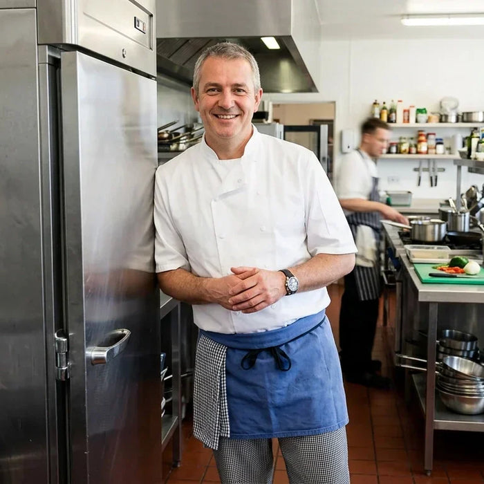 "A professional chef standing next to a stainless steel commercial freezer in a clean commercial kitchen.