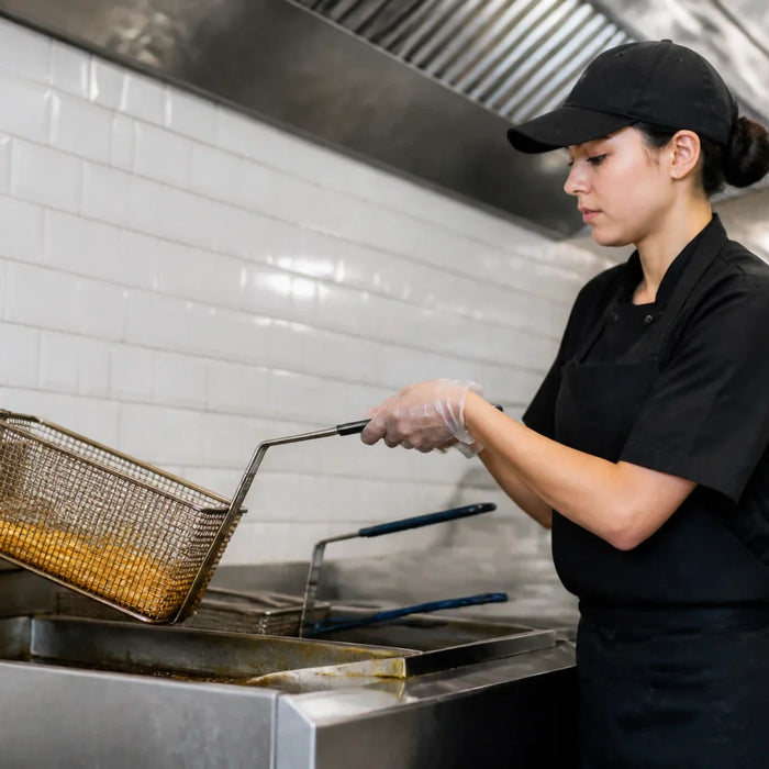 A professional kitchen worker carefully using a stainless steel commercial deep fryer in a fast-casual restaurant setting.