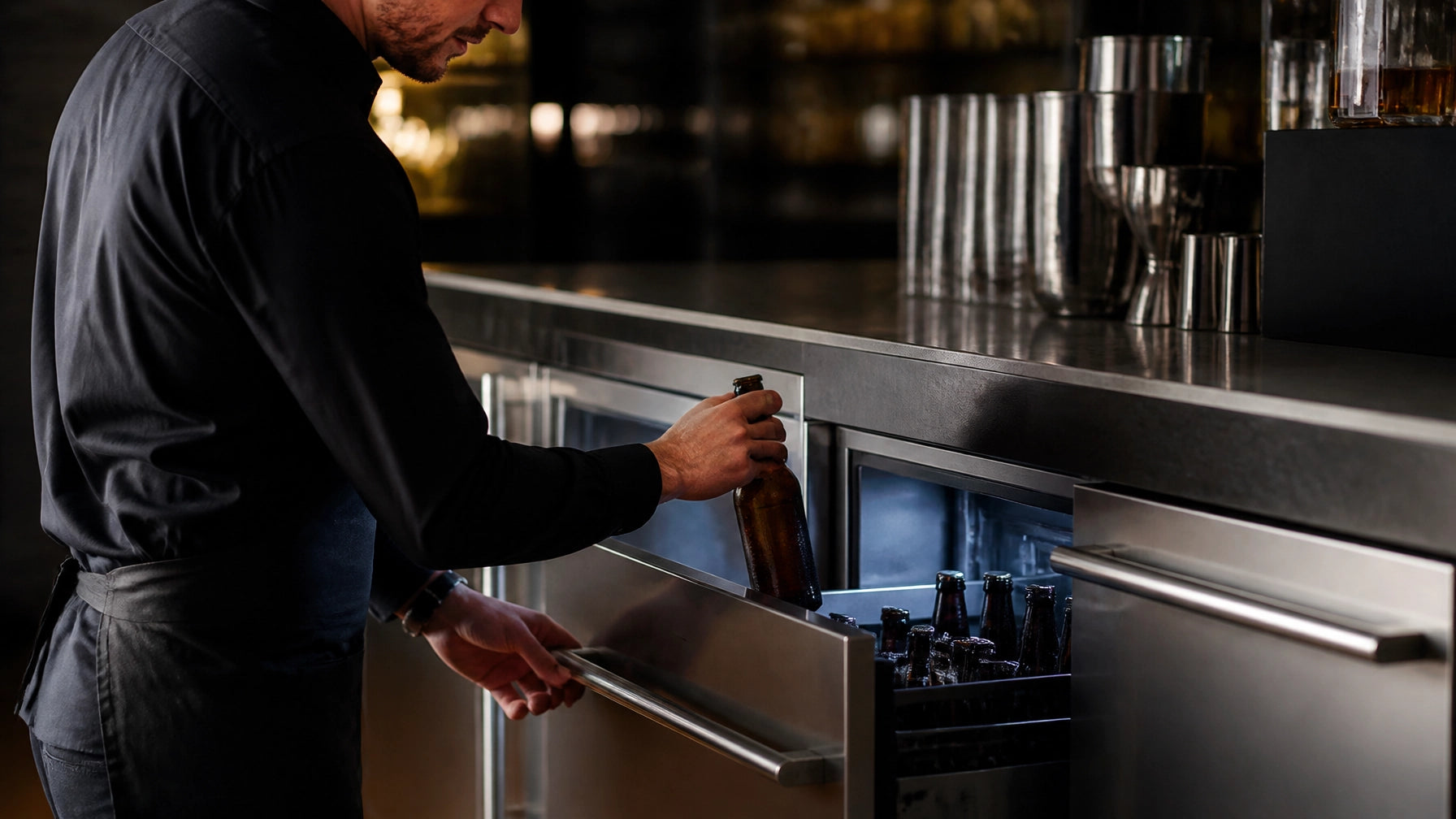 Bartender pulling a bottle from a stainless steel undercounter commercial beer cooler drawer stocked with amber craft beer bottles