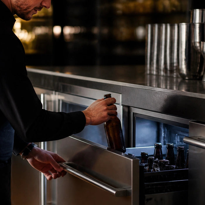 Bartender pulling a bottle from a stainless steel undercounter commercial beer cooler drawer stocked with amber craft beer bottles